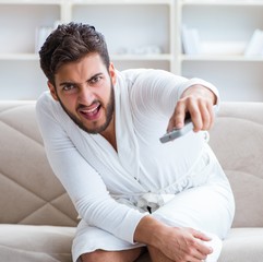 Young man in a bathrobe watching television at home on a sofa co