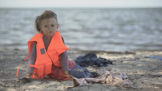 Lonely child refugee in lifejacket sitting shore, dangerous migration across sea