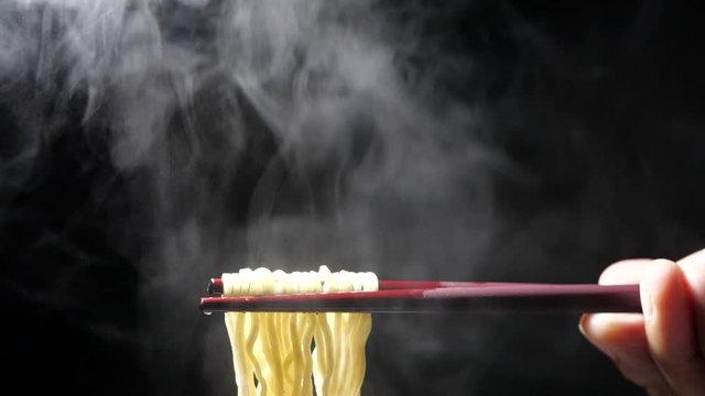 Chopsticks to tasty noodles with steam and smoke in bowl on black background in slow motion, Junk food and fast food concept, Asian meal close-up