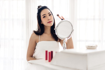 Smiling young beautiful asian woman fresh healthy skin looking on mirror and holding make-up brushes with cosmetics set at home.facial beauty and cosmetic concept