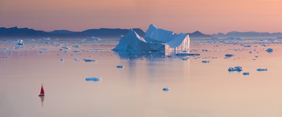Little red sailboat cruising among floating icebergs in Disko Bay glacier during midnight sun season of polar summer. Ilulissat, Greenland. Unesco world heritage © Michal