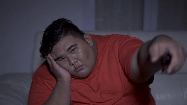 Bored overweight man watching TV on the sofa at night in living room. Shot in 4k resolution