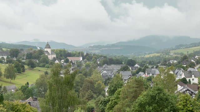 Time lapse of a valley in Sauerland, Germany, with clouds passing by the village of Grafschaft with a clear view of the monastery and typical half-timbered houses against a sloping mountain landscape