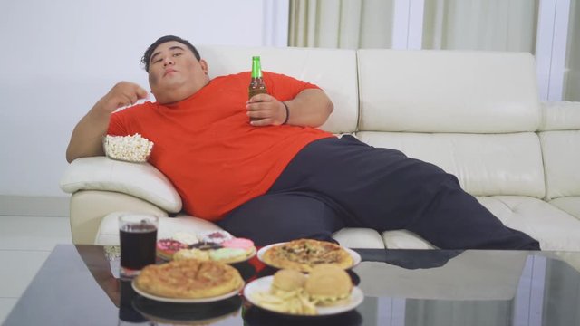 Lazy overweight man enjoying a bottle of beer and junk foods while leaning on the sofa and watching tv in the living room at home. Shot in 4k resolution
