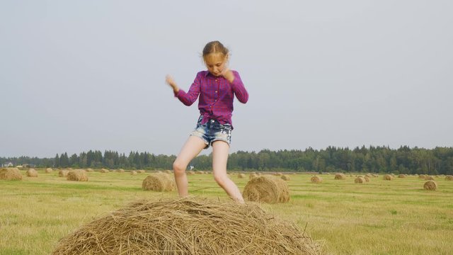 Happy child dancing on haystack. Adorable cheerful pretten girl in checkered shirt and denim shorts dancing on hay in autumn field. Harvest concept