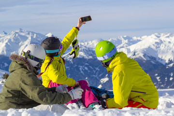 Three happy friends snowboarders and skier are having lunchtime and make selfie on a smartphone on a ski slope in sunny day in the mountains.