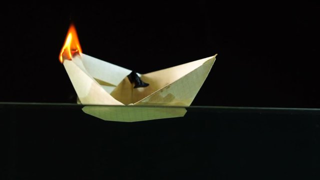 A paper boat burns with fire and floats in water on a black background, the concept of tragedy, slow motion, close-up