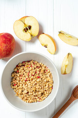 white wheat granola in a white bowl in a composition with a spoon and apple on white wooden background. Healthy breakfast food. Making breakfast. Gluten free