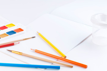 Side view of an open drawing album with blank pages, bright watercolor paints, paint brushes and colored pencils on a white table