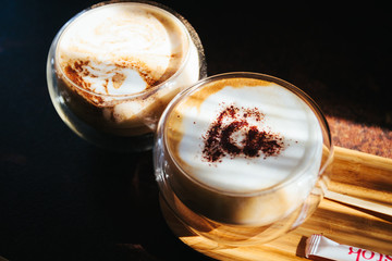 Cappuccino in a transparent glass in the morning sun. Morning in cafeteria. Sunlight. Close up. Coffee mood. Light cup in coffee shop. Sweet and tasty latter macchiato in flare cup on table.