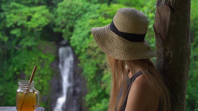 Slowmotion shot of a young woman in a beautiful cafe with an epic view on a canyon and waterfall drinks tea with ice. Travell to Bali concept