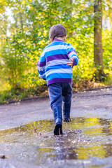 Obraz premium A little boy is jumping in a puddle. A boy in rubber boots. Happy childhood. Puddles after the rain. Warm summer evening.