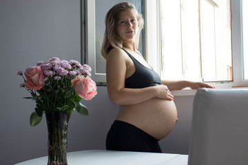 Pregnant woman in a beautiful colorful dress is standing next to a bright bouquet of flowers and holds hands on belly at home interiors. Pregnancy, parenthood, preparation and expectation concept