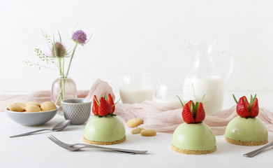 White wooden table with 3 no-bake strawberry mini sphere matcha cheesecakes with bowl of salt caramel, buiscuits, glass pitcher and glass of milk and glass vase with wild teasel flowers.