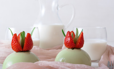 Matcha cheesecakes. White wooden table with 2 no-bake strawberry mini sphere cheesecakes embellished with real fresh strawberries and leaves of rosemary with glass pitches and glass of milk.