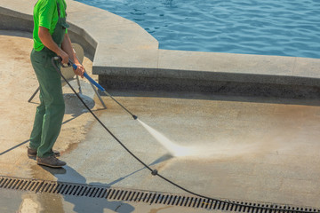 Worker man in uniform washes street or park sidewalk near water pool or fountain. Municipal service...