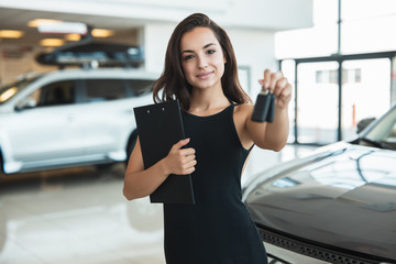 beautiful brunette woman manager holding brochure in one hand and car keys in another standing in dealership center