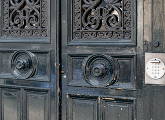 Old wooden door. Weathered double door of black painted wooden framed panels with black casting metal ornate gratings and circle doorknobs. Metal intercom button panel on wall of old building 