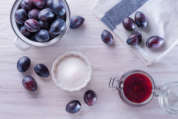 An enamel pan with fresh mature plums, an open glass jar with homemade plum jam, folded towel and white ceramic bowl with sugar on a wooden table. Flat lay, top view