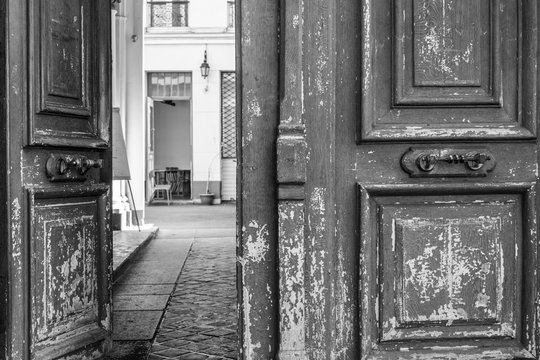 Old wooden door. Black and white photo of shabby double door surface with peeling paint. Opened door to patio inside old house in Paris France. Vintage framed wooden door details. City life scene. 