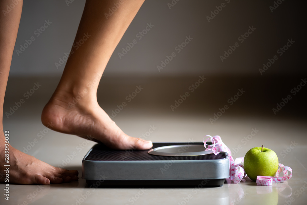 Wall mural Female leg stepping on weigh scales with measuring tape and green apple. Healthy lifestyle, food and sport concept.