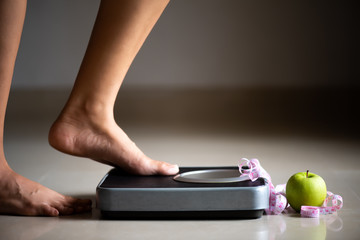 Female leg stepping on weigh scales with measuring tape and green apple. Healthy lifestyle, food...