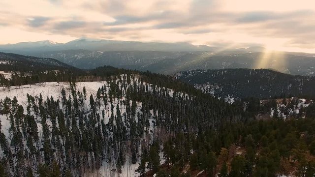 Drone flies above snowy forest landscape towards rays of sun through clouds.