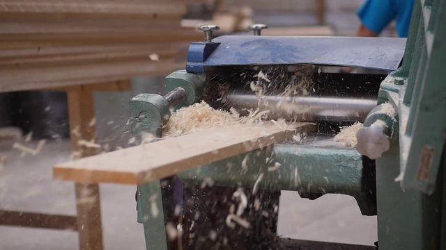 A solid wood plank coming out from an old planing machine. Slow motion shot of the plane tool processing wood while wood shavings are flying towards the camera. Shot taken in a woodworking workshop.