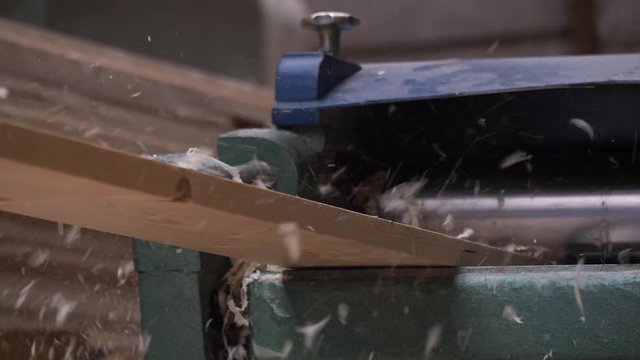 A solid wood plank coming out from an old planing machine. Slow motion shot of the plane tool processing wood while wood shavings are flying towards the camera. Shot taken in a woodworking workshop.