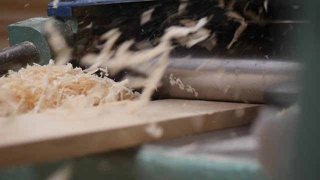 A solid wood plank coming out from an old planing machine. Slow motion shot of the plane tool processing wood while wood shavings are flying towards the camera. Shot taken in a woodworking workshop.