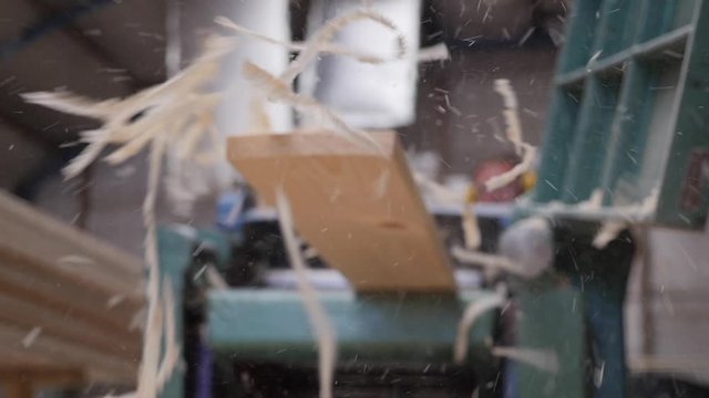 A solid wood plank coming out from an old planing machine. Slow motion shot of the plane tool processing wood while wood shavings are flying towards the camera. Shot taken in a woodworking workshop.