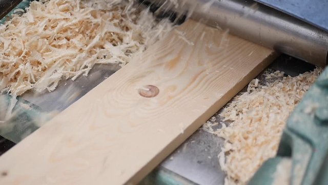 A solid wood plank coming out from an old planing machine. Slow motion shot of the plane tool processing wood while wood shavings are flying towards the camera. Shot taken in a woodworking workshop.
