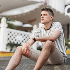 Fashionable European young man in a stylish striped t-shirt in summer beige shorts sits on the...