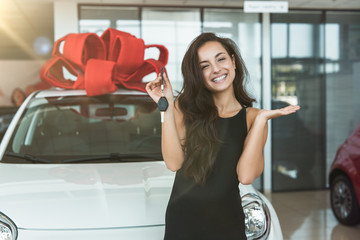 young beautiful woman feels happy holding keys from her brand new car standing in dealership center