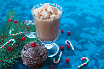  Christmas homemade chocolate muffin decorated with red berries and a glass of hot cocoa on a background of a Christmas tree branch.