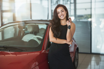 young beautiful woman looks happy with car keys in her hand standing in dealership center buying brand new car