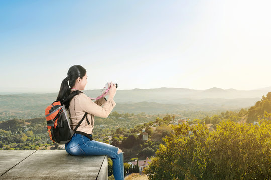Asian woman with a backpack sitting on the rooftop and holding a camera to take pictures