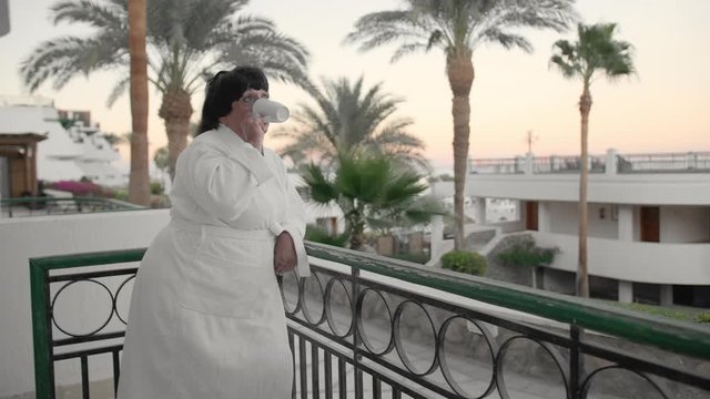 Caucasian senior woman in the morning drinking coffee on the hotel terrace in a white terry robe at a tropical resort. Against the backdrop of palm trees and the sea. Slow Motion.