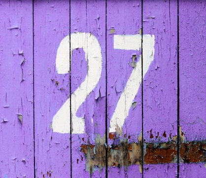 Arabic figure 27 (twenty seven) painted with white on an old wooden wall covered with violet heavily peeled paint