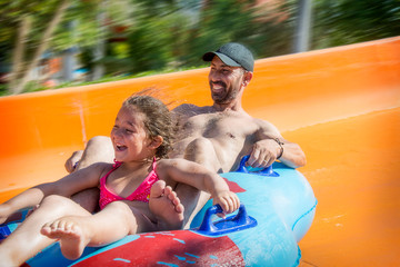 In the summer, in a water park, a girl and dad ride down the hill on an air mattress.