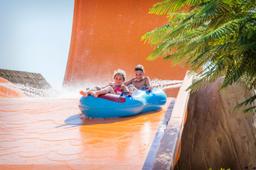 In the summer, on a bright sunny day, in a water park, a brother and sister ride off a slide on an air mattress.