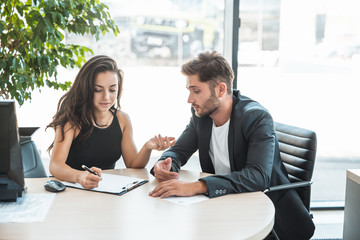 beautiful young woman manager assisting her handsome man client fulfilling papers during purchase...