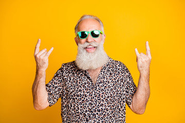Portrait of cheerful funky funny old bearded man with eyewear eyeglasses showing horns sign enjoy rock and rool party event wearing leopard print shirt isolated over yellow background