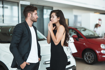 beautiful young woman trying to persuade her handsome husband to buy a new car standing in dealership center