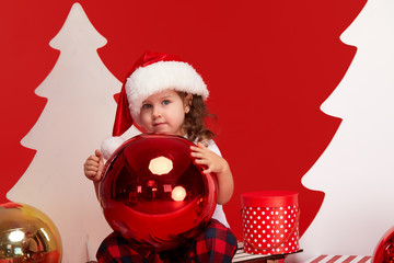 Little child girl in santa claus hat sits on wooden sleigh with gift boxes and holds big christmas ball on red isolated background. blank space for text. Little Santa helper