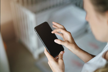 Top view, close-up is smartphone in female hands. In background crib in soft focus. Mom businesswoman works remotely.