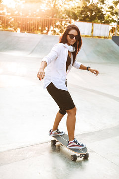 Photo of beautiful young woman wearing headphones riding skateboard in skate park
