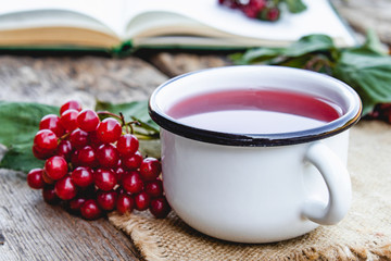 White mug or cup of hot viburnum tea on a wooden table near an open book and red viburnum berries. Source of natural vitamins. Used in folk medicine.