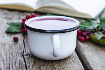 White mug or cup of hot viburnum tea on a wooden table near an open book and red viburnum berries. Source of natural vitamins. Used in folk medicine.