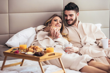 Young happy couple having breakfast in luxury hotel room, watching TV.